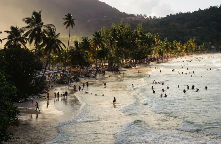 The rays from the setting sun filter through palm trees as locals enjoy the surf at Maracas Beach, Trinidad, Trinidad & Tobago